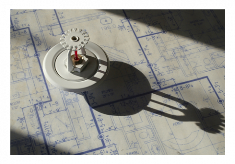 A small mechanical model with gears, casting a shadow, placed on top of a blueprint. The blueprint displays detailed architectural or engineering plans with various measurements and schematic symbols. The model appears to be metallic and intricate in design.