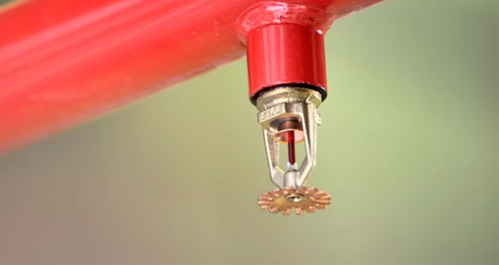 Close-up of a fire sprinkler head mounted on a red pipe. The sprinkler consists of a metal frame holding a heat-sensitive element designed to activate when temperatures rise, allowing water to spray through the circular deflector for fire suppression. Background is blurred.